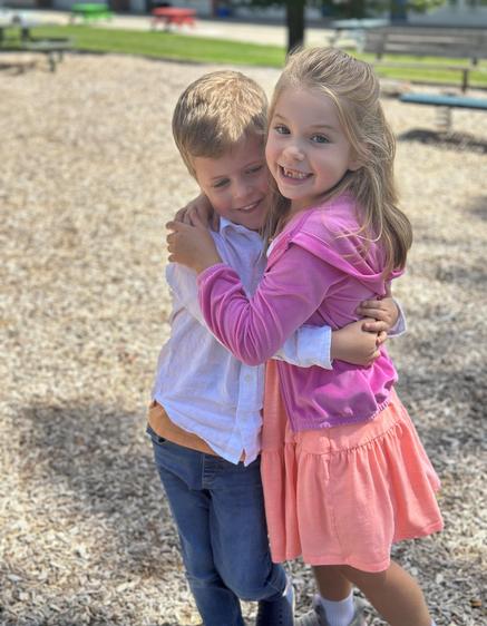 Two children embrace happily on a playground covered in wood chips.