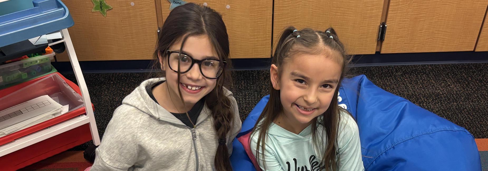 Two girls in classroom reading and smiling.