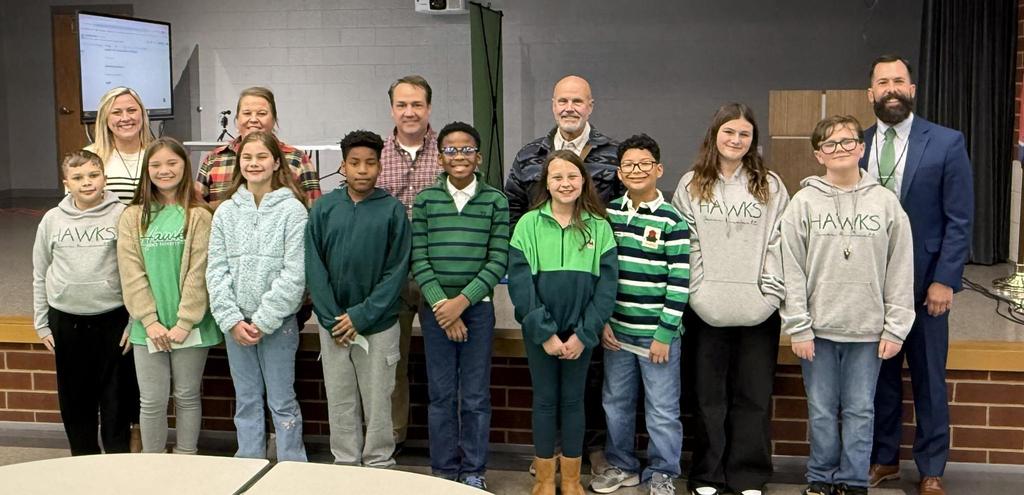 9 kids dressed in greens and blues standing in front of 5 adults standing in front of a stage smiling