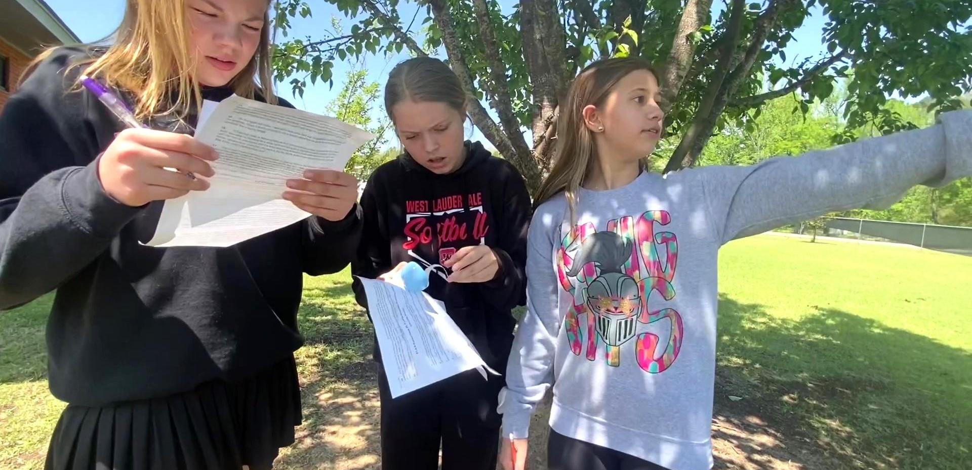 Three girls studying and pointing outdoors, focusing on their papers.