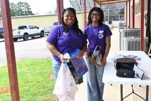 Two women in purple shirts smiling while holding bags and standing outside.