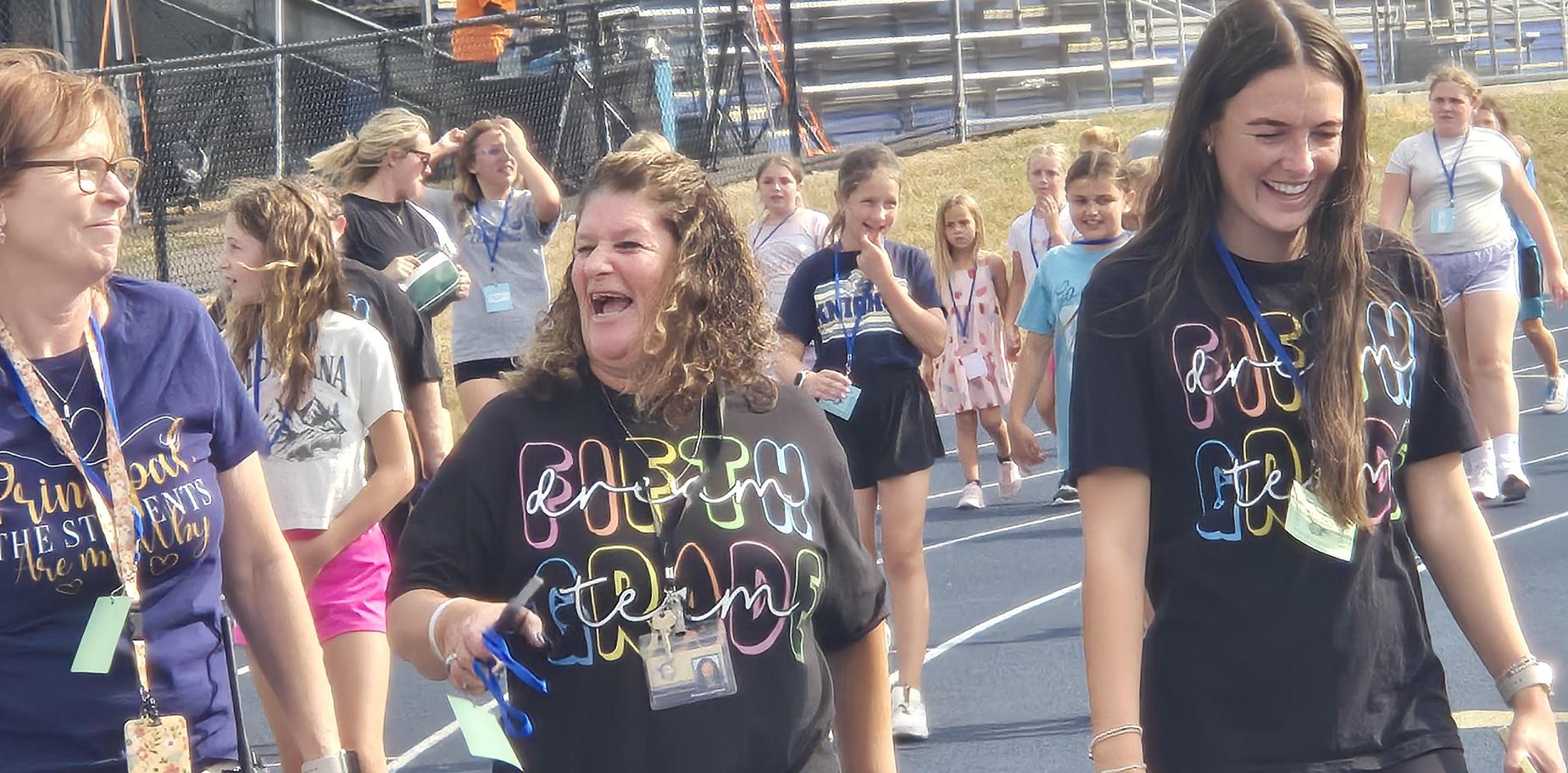 Smiling women in matching shirts engaging with children as they walk on a track.