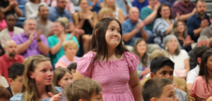 Girl in pink dress stands while surrounded by classmates and audience