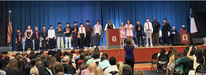 Students stand on stage with red medals as their coach speaks at the podium
