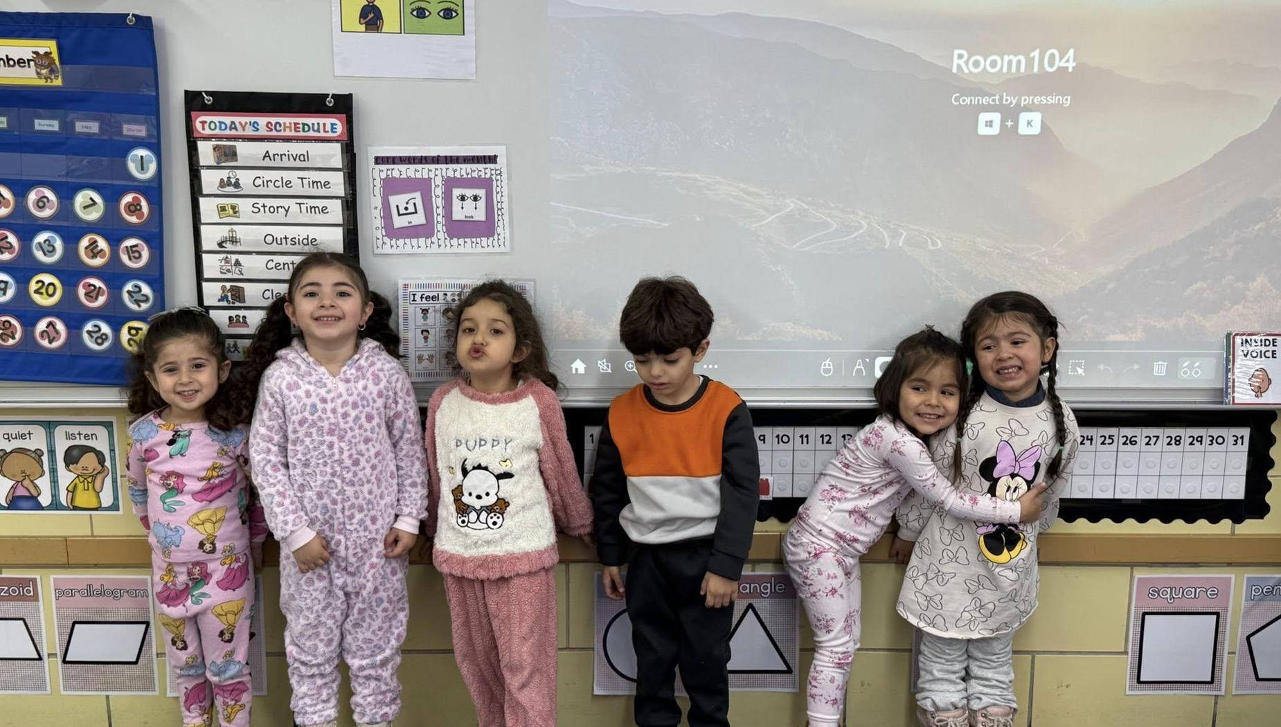 Seven kids in pajamas standing in front of a classroom screen, smiling happily.