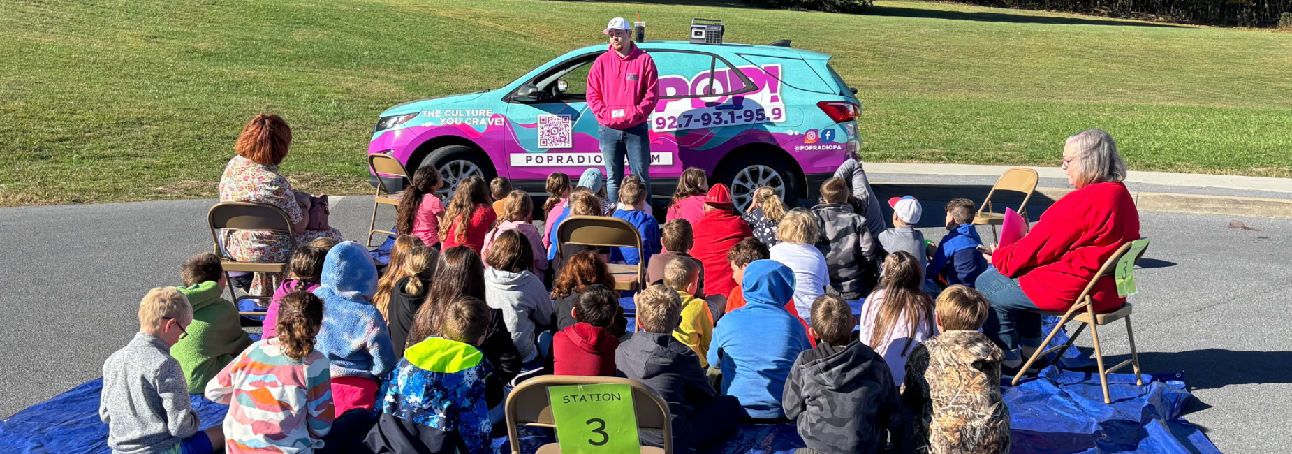 Kids seated on chairs and a blanket, engaged with a speaker beside a colorful vehicle.