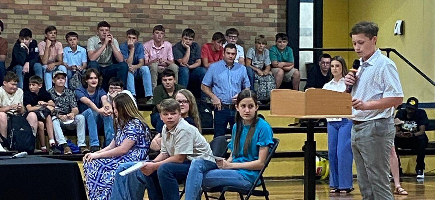 A group of students seated in an assembly, with one speaking at a podium.
