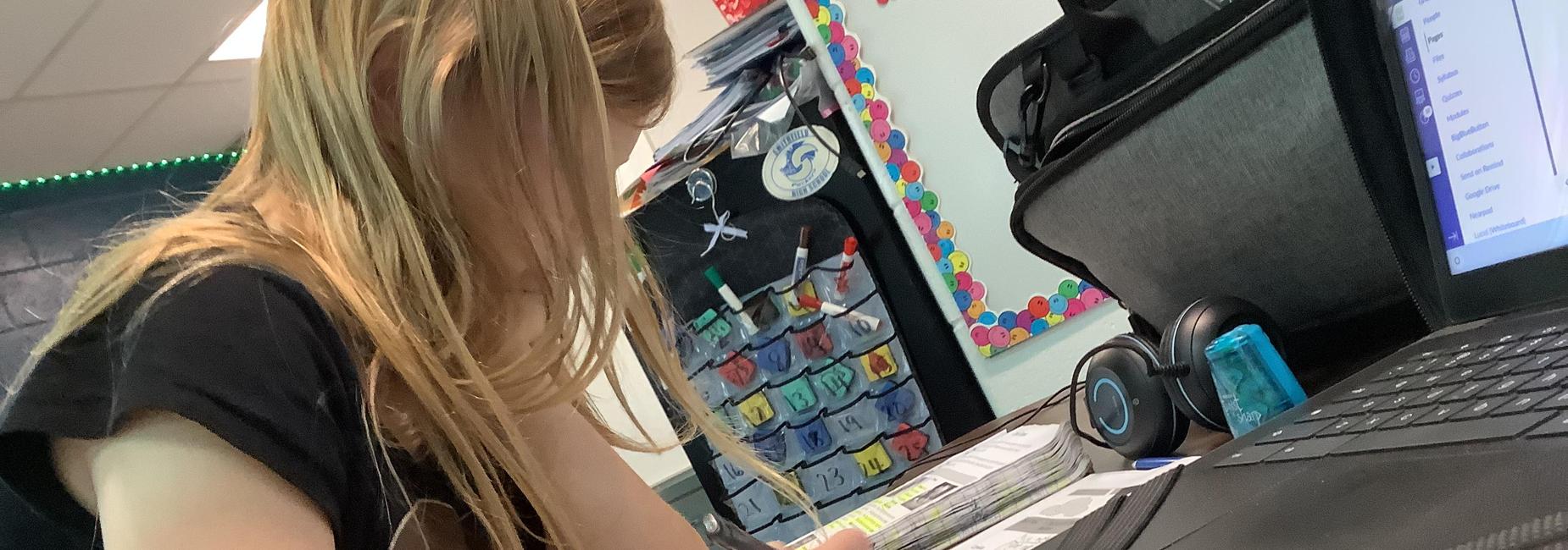 A student is writing at a desk with a laptop and colorful calendar in the background.