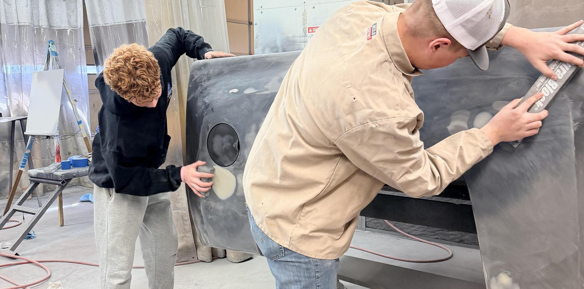 Two individuals sanding a car body in a workshop.