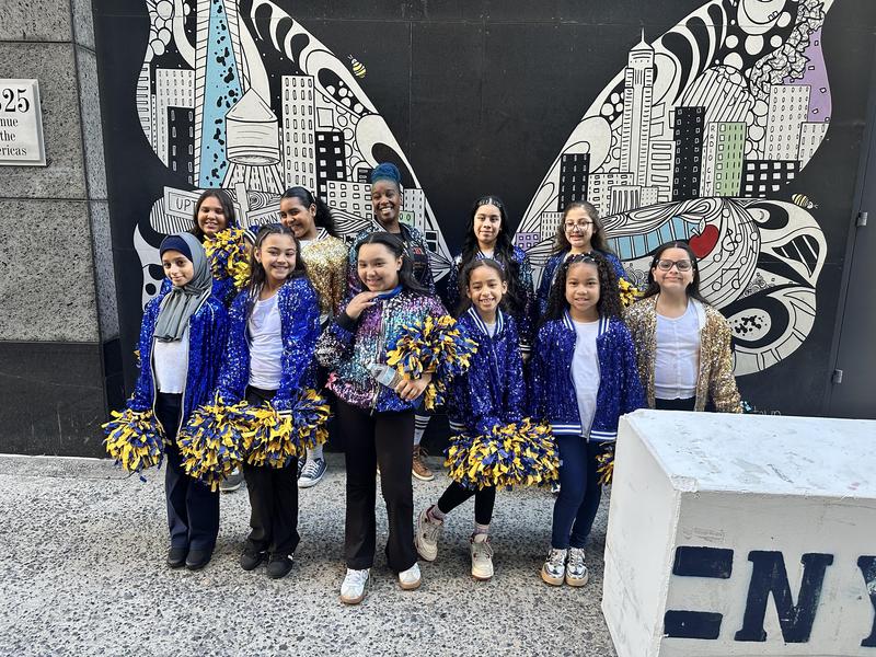 A group of cheerleaders in sparkly jackets poses in front of a colorful mural.
