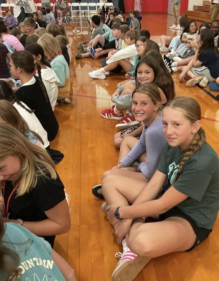 A group of children sitting on a gym floor, smiling towards the camera.