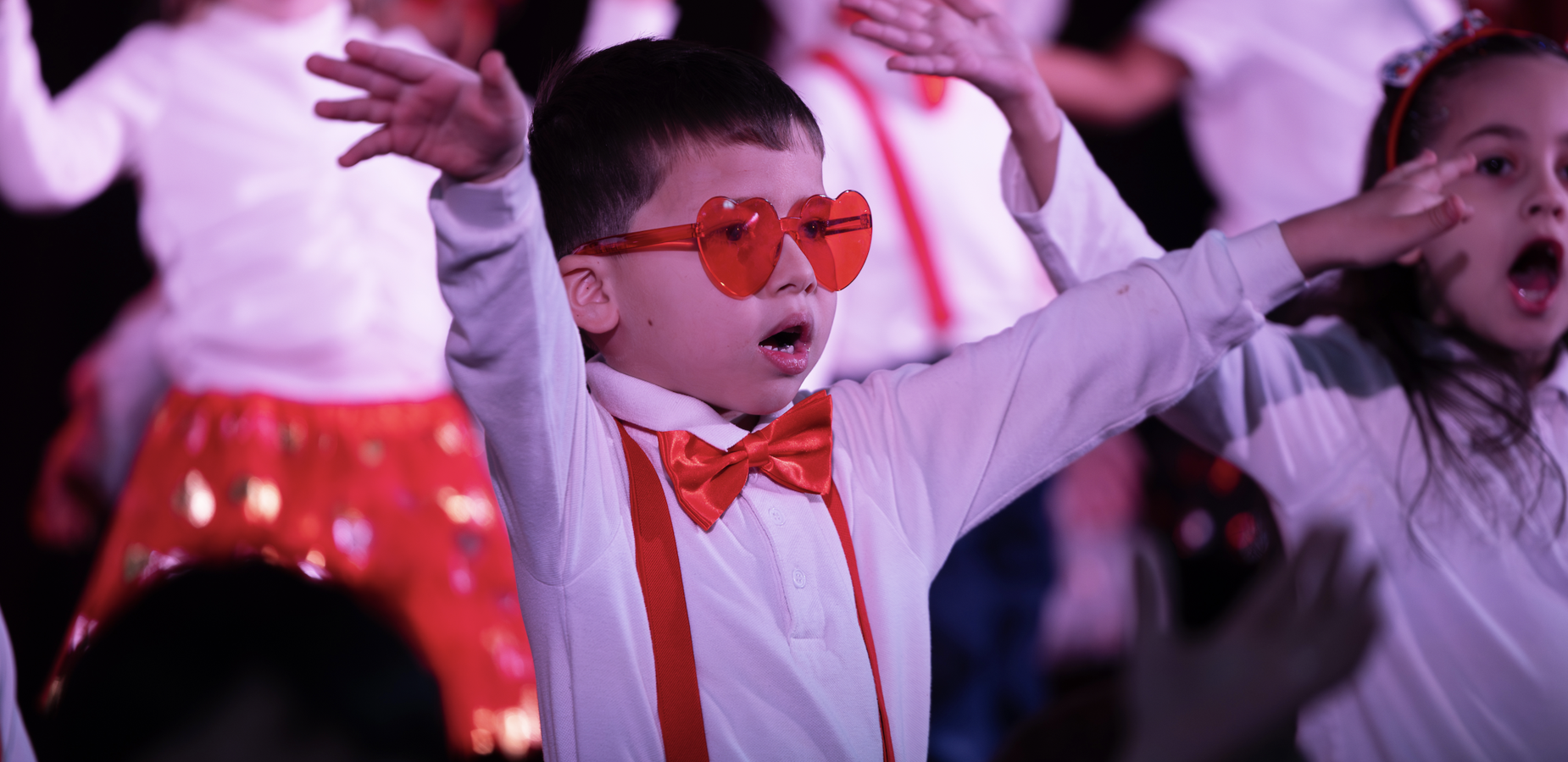 Child in heart-shaped sunglasses performing on stage with other children.