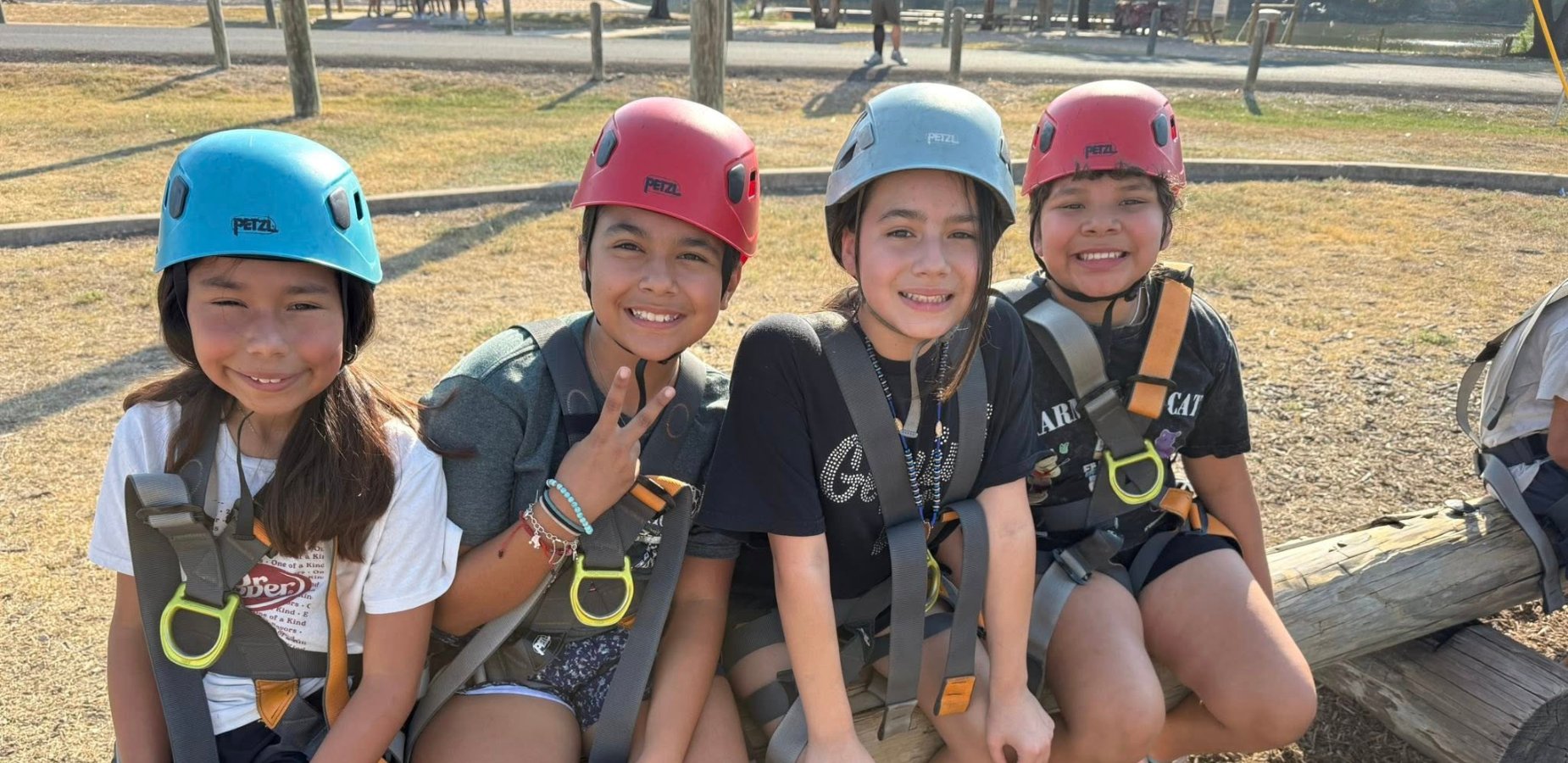 4 girls wearing blue or red helmets and harnesses for zipline, sitting on a log