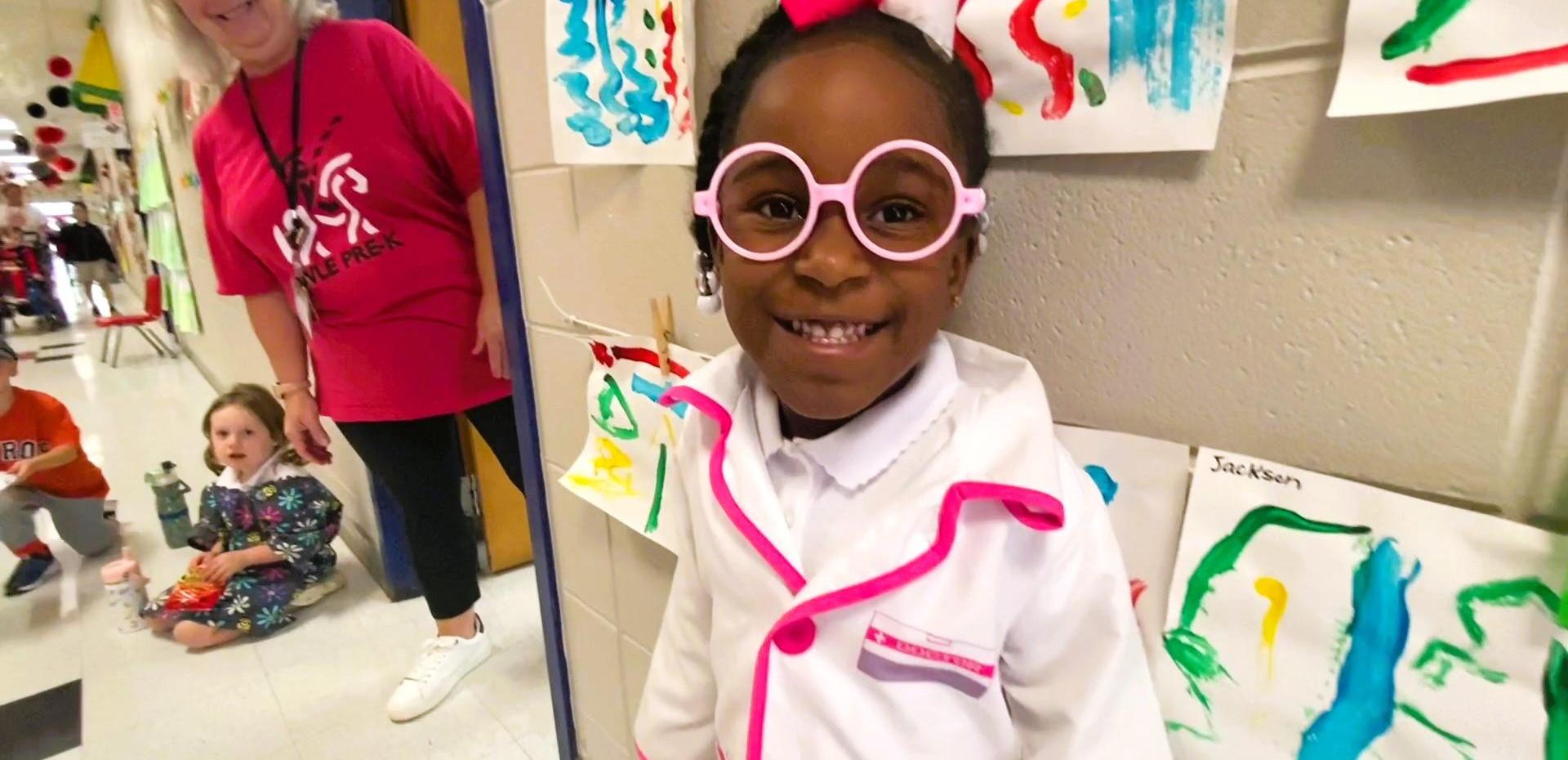 Smiling girl in glasses wearing a lab coat, surrounded by colorful artwork on the walls.
