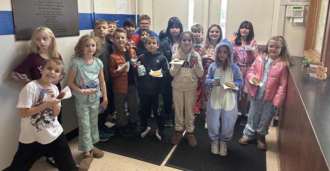 A group of children in pajamas holding snacks and drinks in a school hallway.