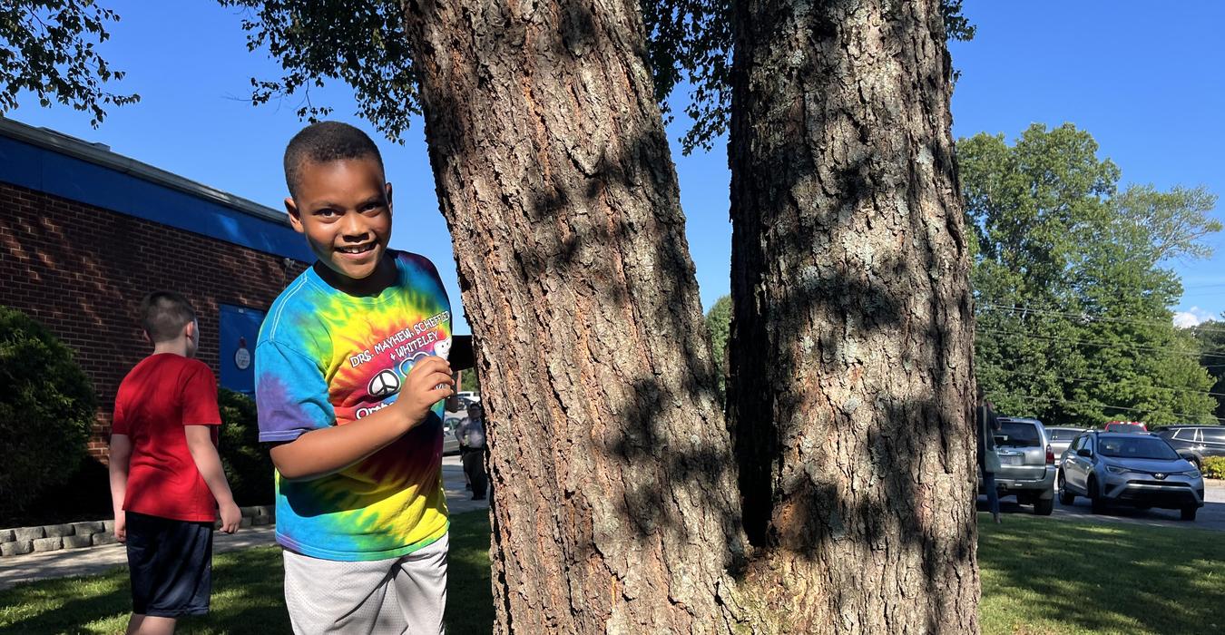 A child smiling beside a large tree in an outdoor setting.