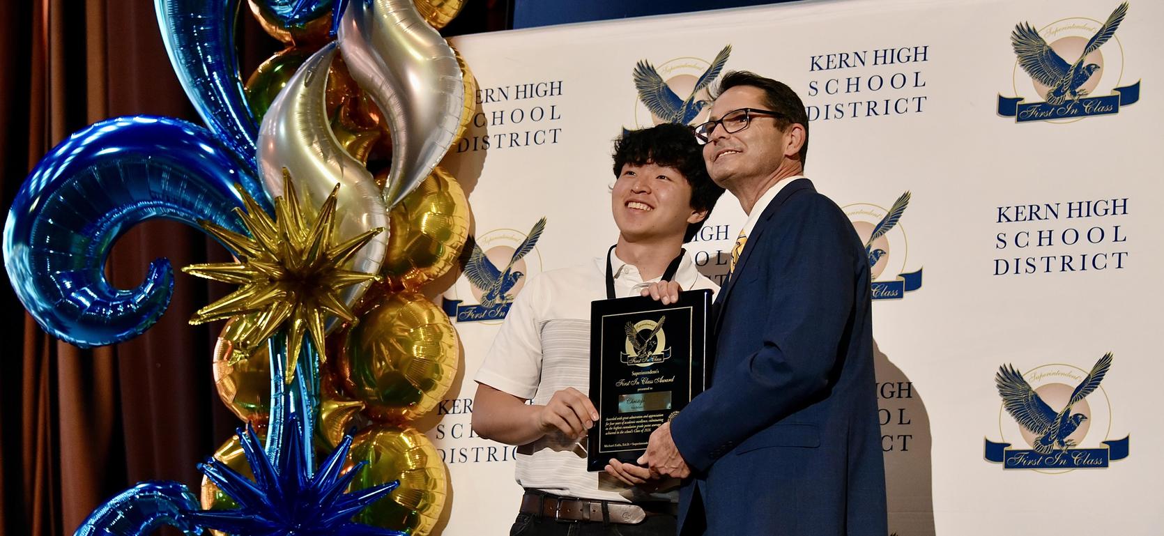 Two individuals holding an award in front of a decorated backdrop.