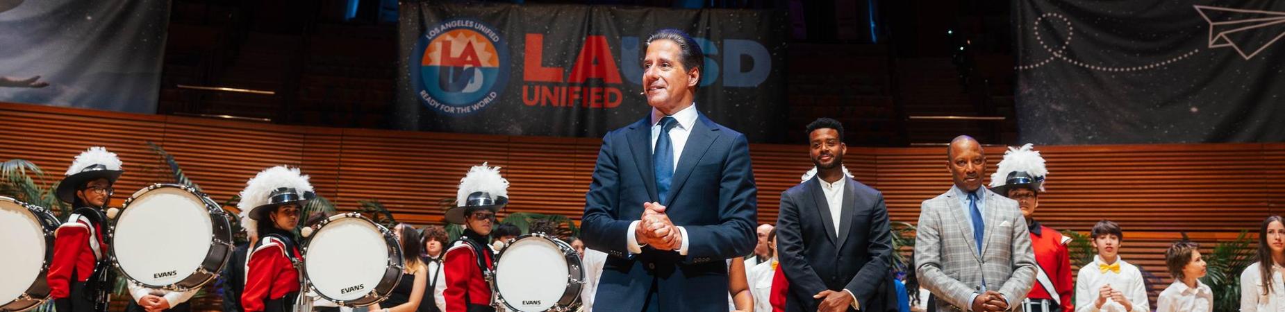 A group of people standing on a stage with a large LAUSD Unified screen in the background. Several individuals in uniforms hold musical instruments.