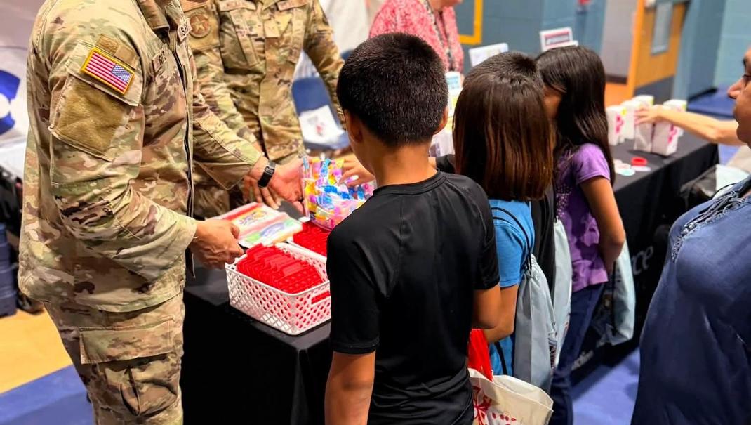 Children gather around a table with military personnel organizing school supplies.