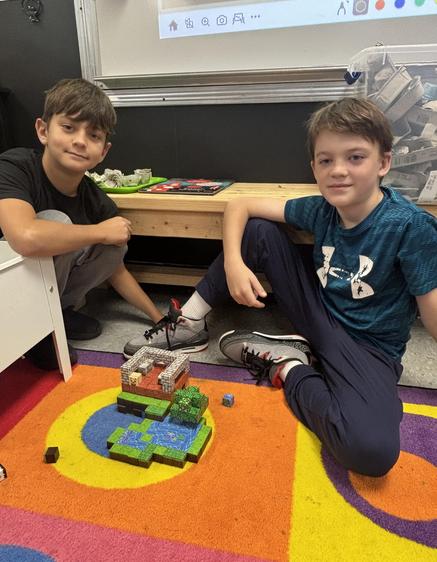 Two boys sit on the floor with a Lego creation, smiling at the camera.