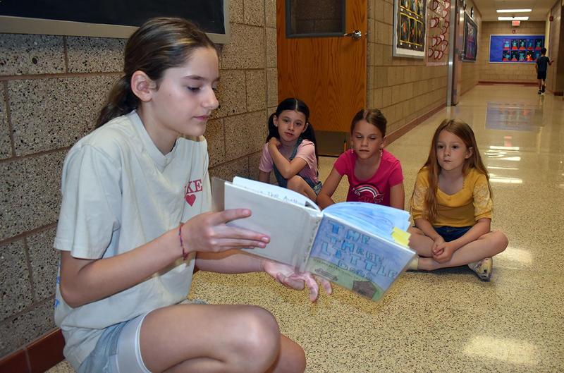 A girl reads a book to three seated classmates in a school hallway.