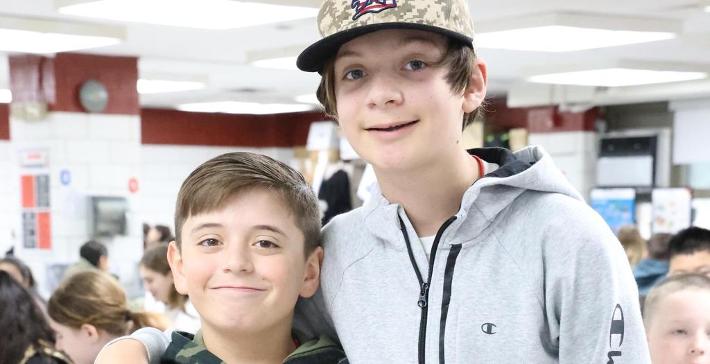 A tall boy with his arm around a boy wearing camouflage clothing.