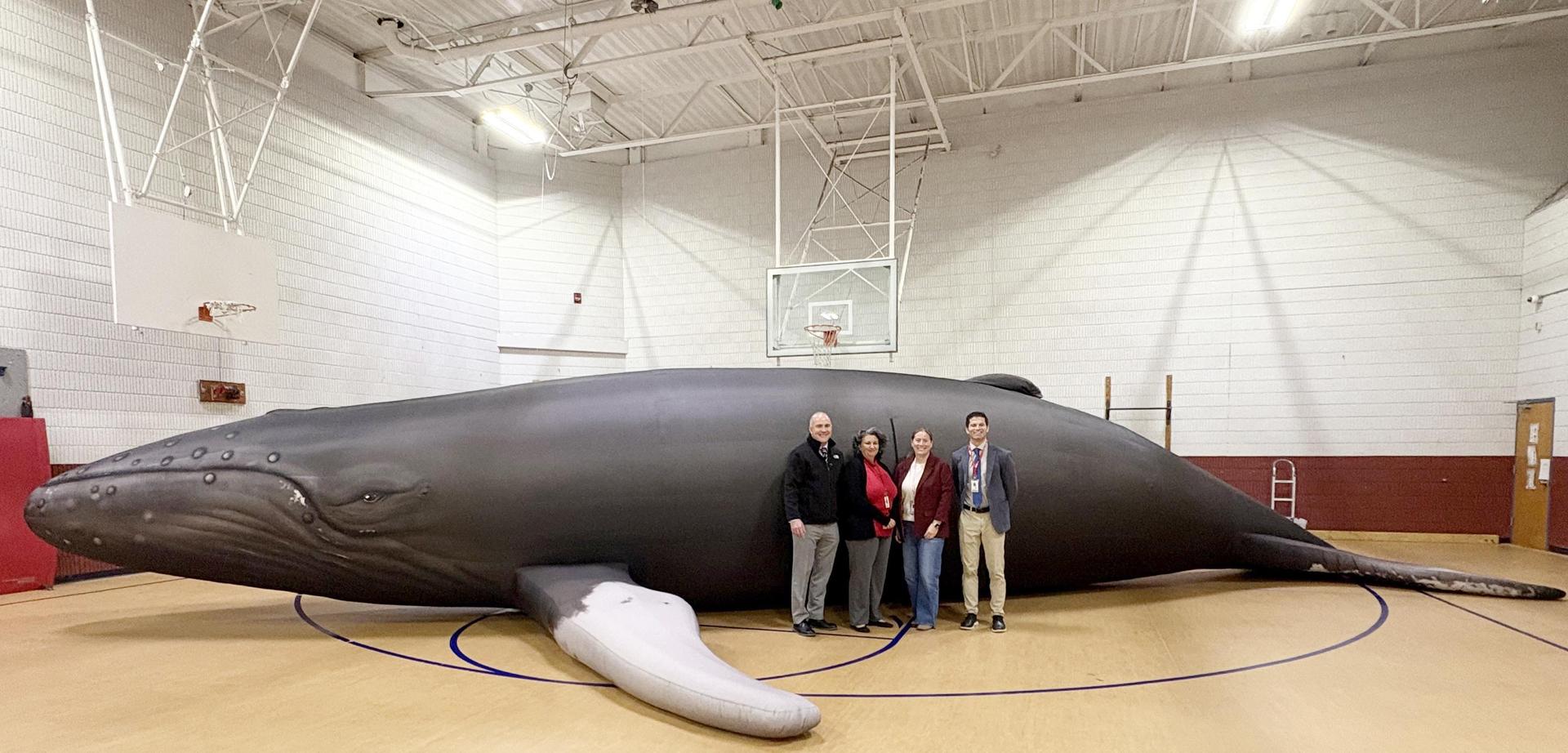 Adults standing beside a large inflatable whale in a gymnasium.