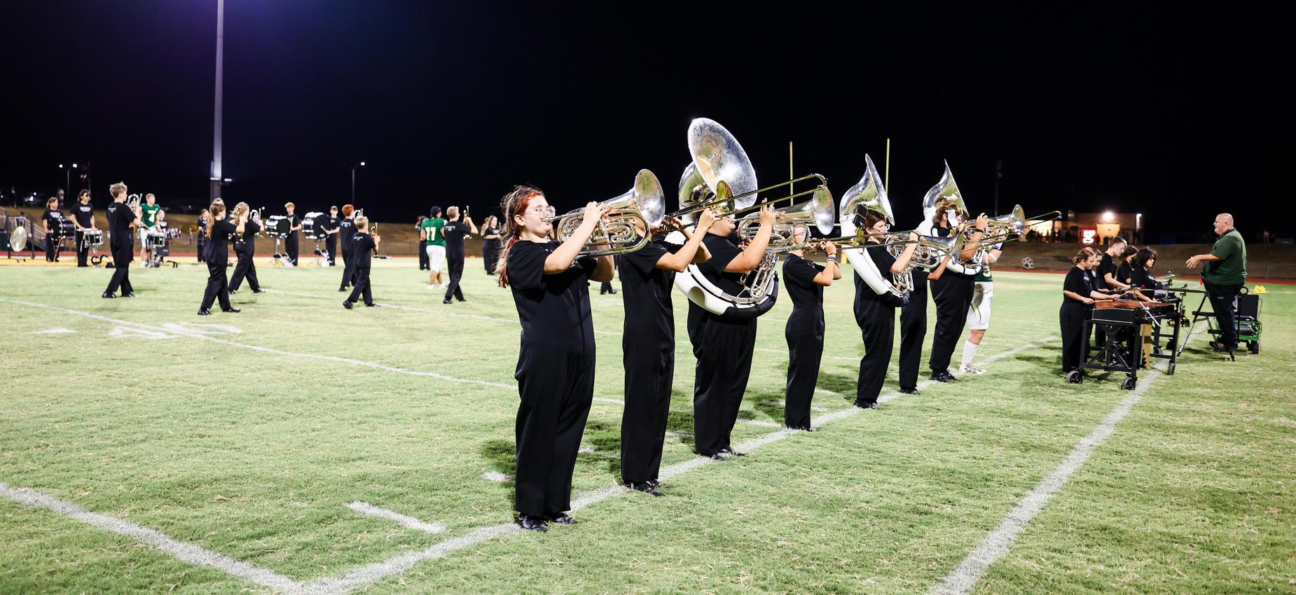 Marching band members playing brass instruments on a football field at night.