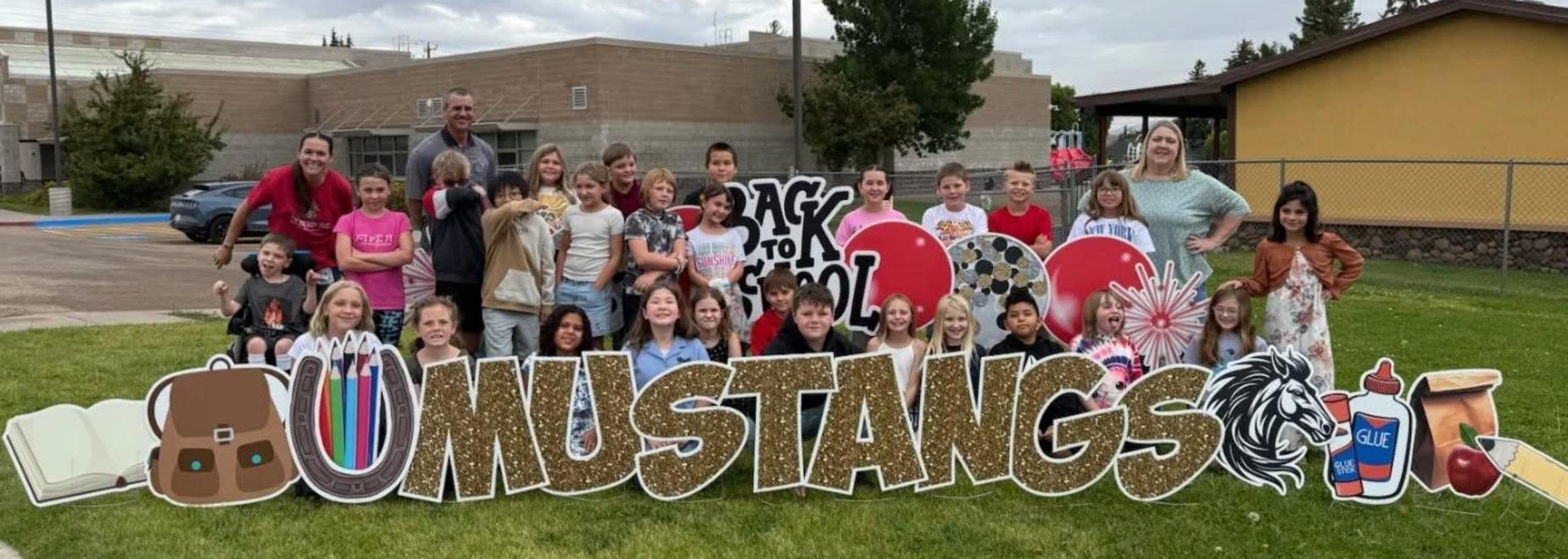 Another group of children gathered around a colorful 'Mustangs' school sign outdoors.