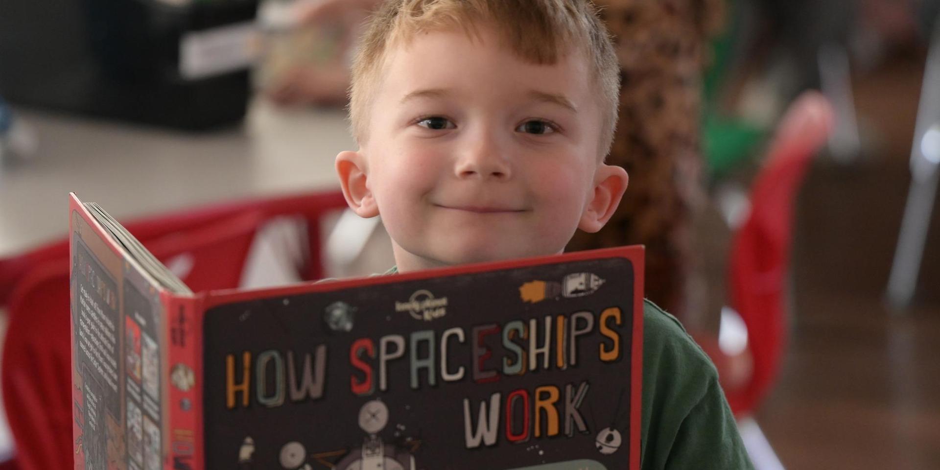 A boy holding a book titled 'How Spaceships Work' with a smile.
