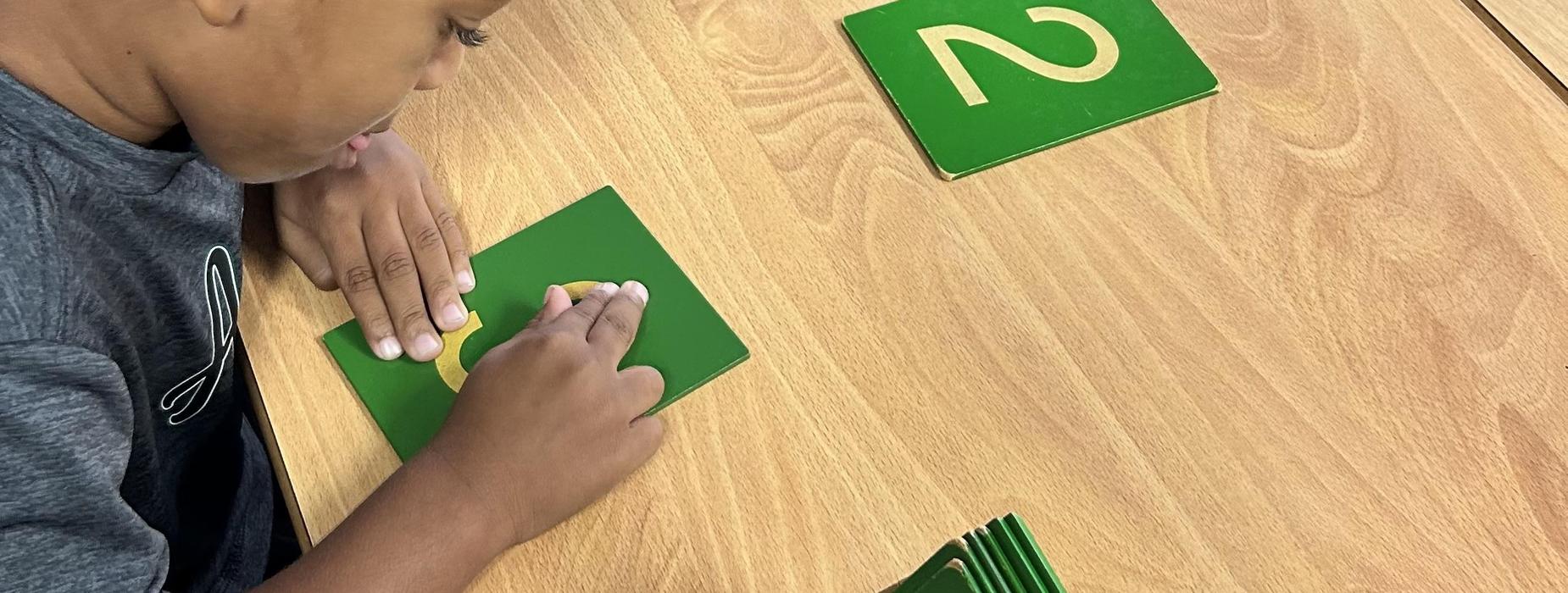 A child tracing shapes on green cards at a wooden table in a classroom setting.