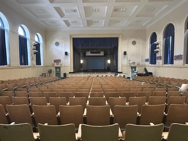 An empty auditorium with rows of seats facing a stage and large windows.