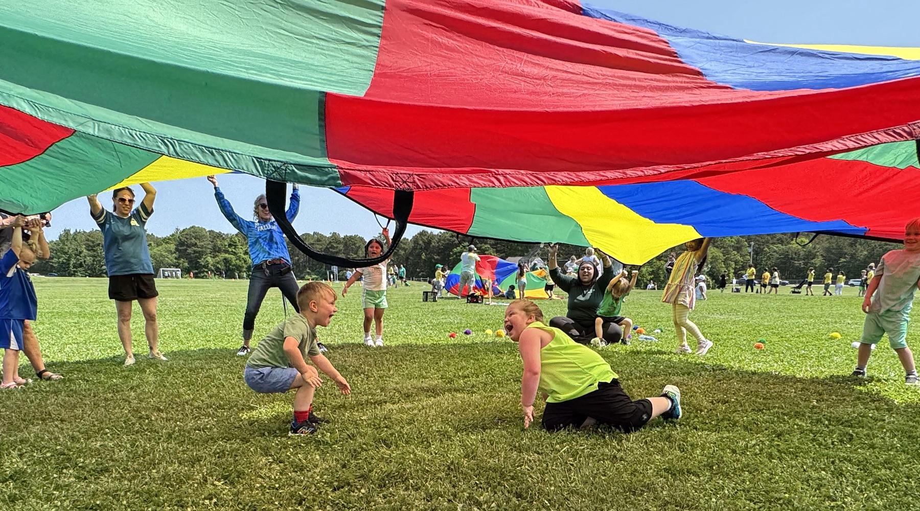 Children play under a large colorful parachute in a grassy field.