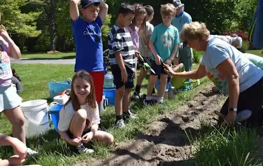 Children gathered around an adult, learning to plant seeds in a garden setting.