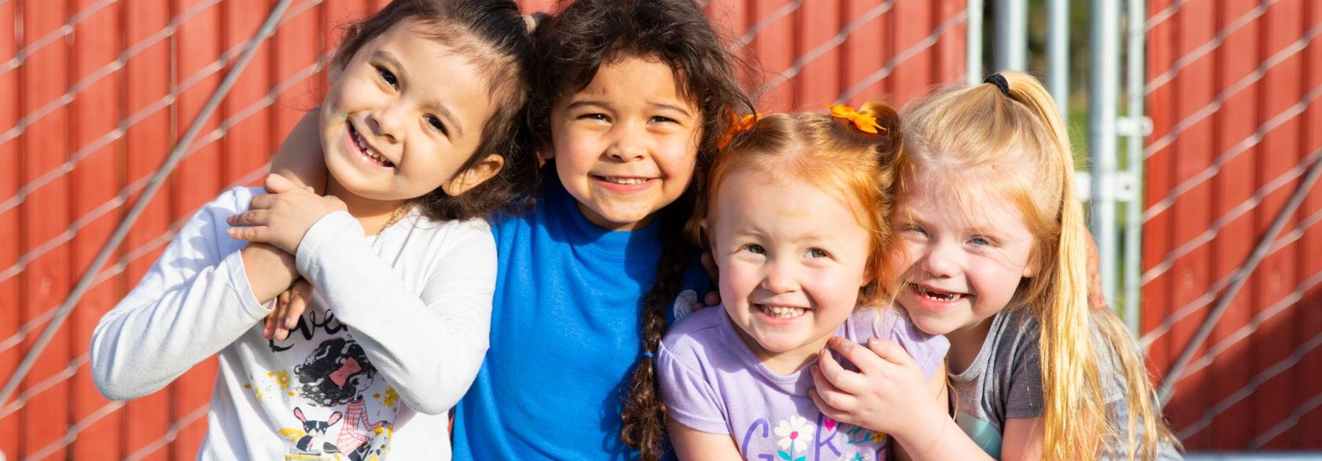Four young children smile and sit together outdoors in front of a playground structure.