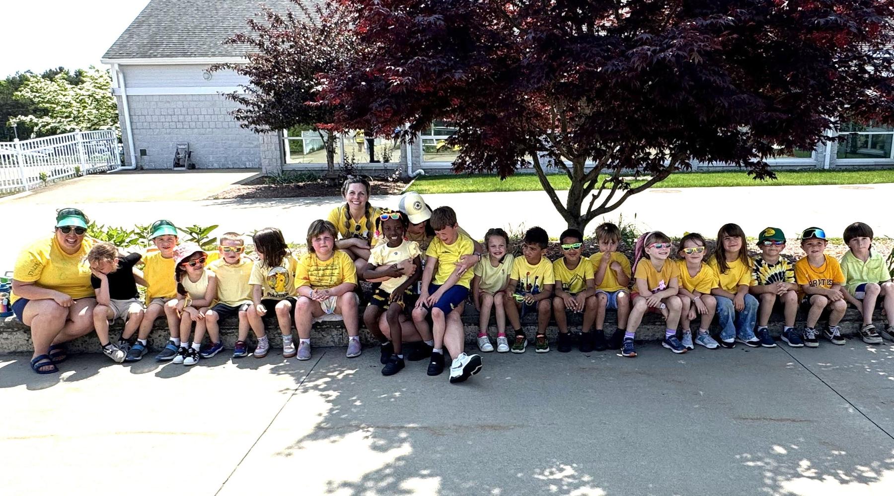 A group of children in yellow shirts gathered under a tree for a photo.
