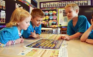 Three children in blue shirts examine a colorful chart at a classroom table.