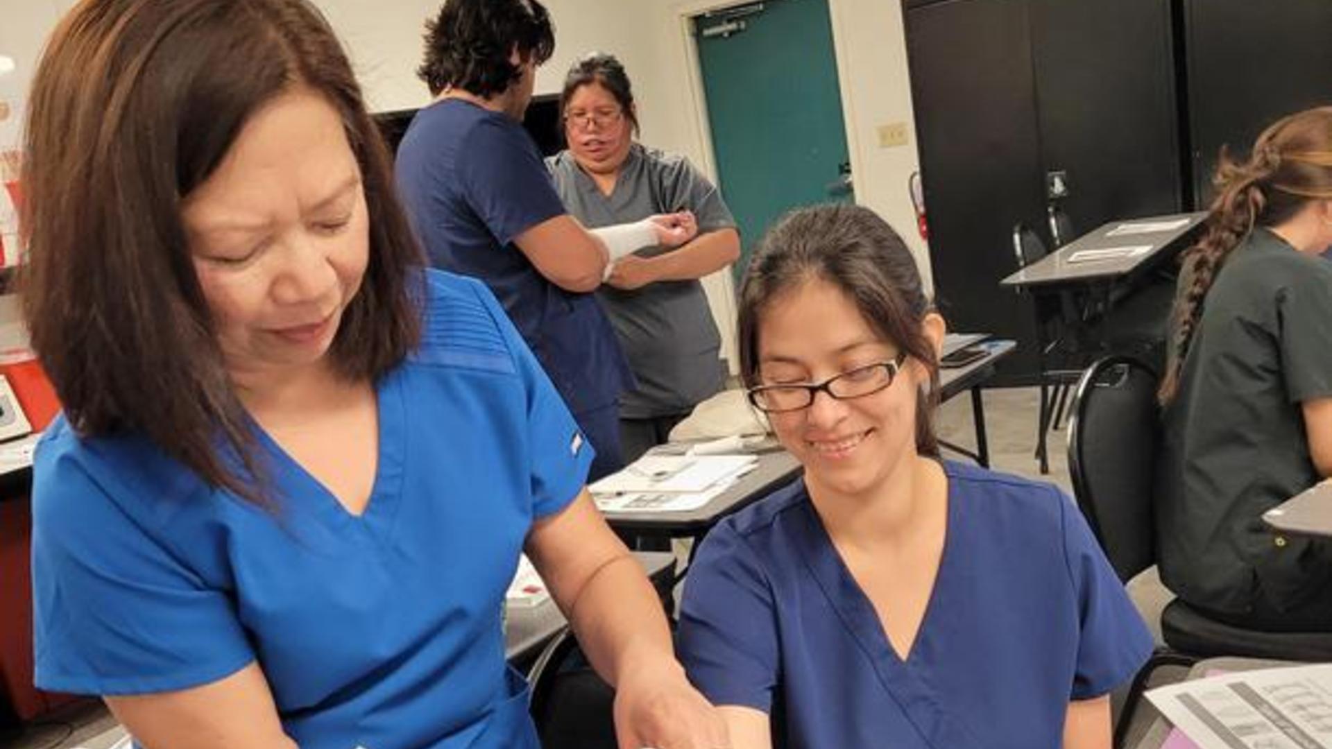 Adult students in blue and gray scrubs practicing first aid skills in a classroom