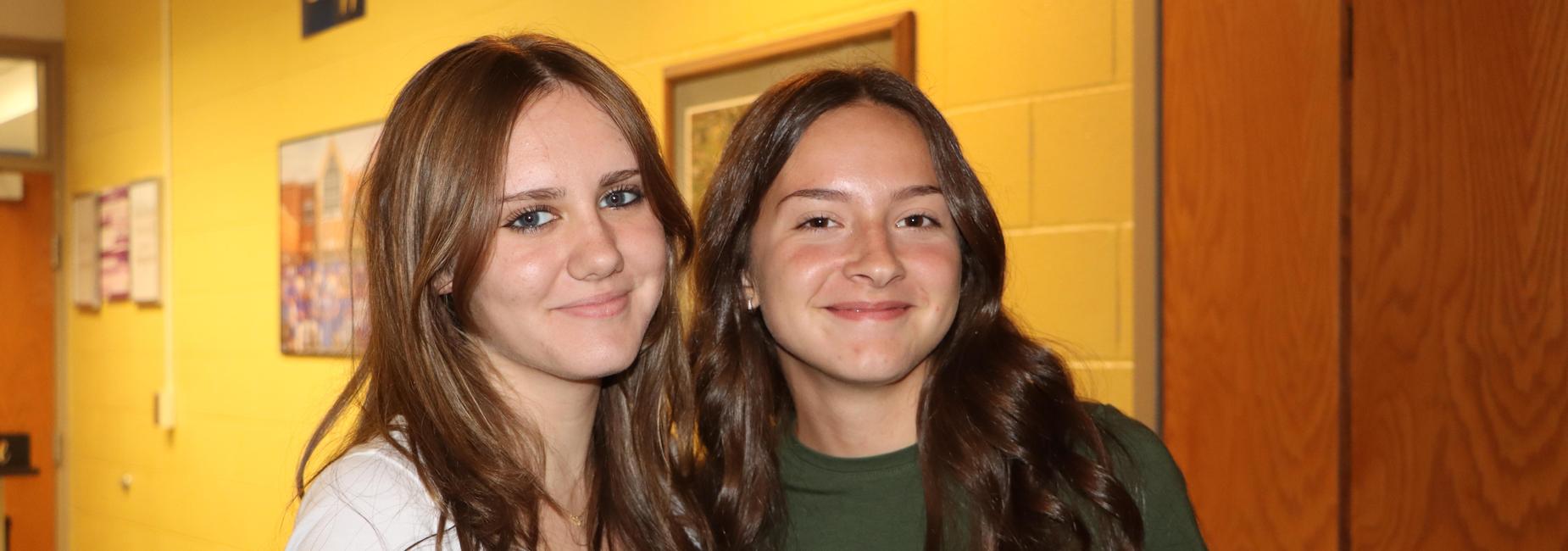 Two smiling teenage girls posing together in a hallway with yellow walls.
