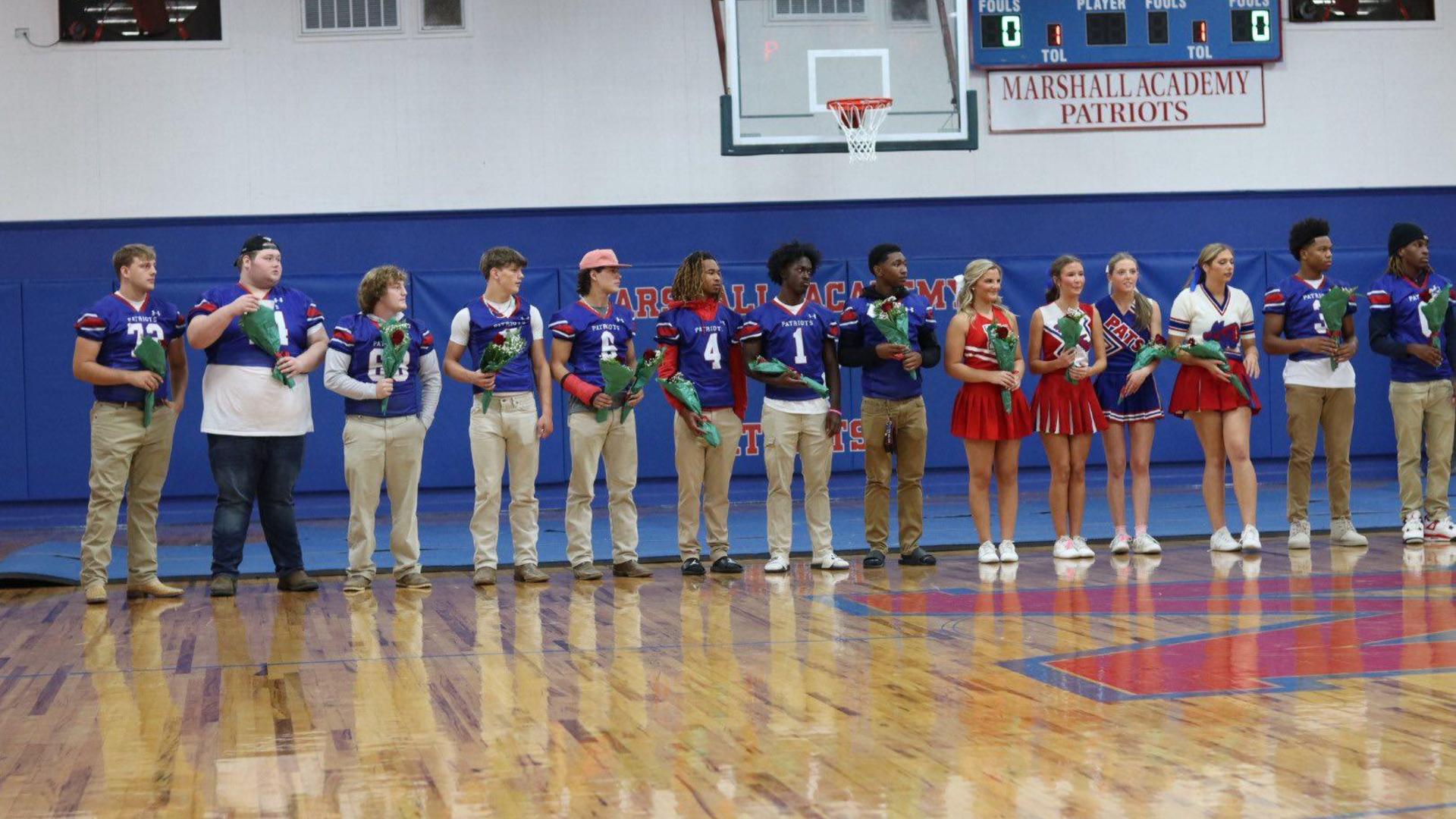 Students in football jerseys and cheerleading uniforms hold roses on a gym floor.