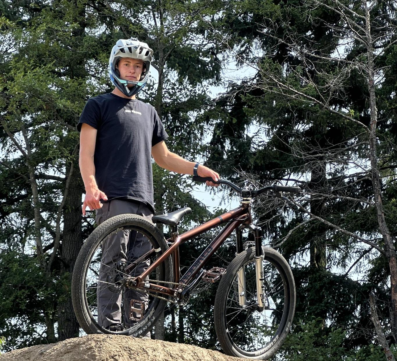 Young person wearing a helmet stands on a bike on a dirt mound surrounded by trees.
