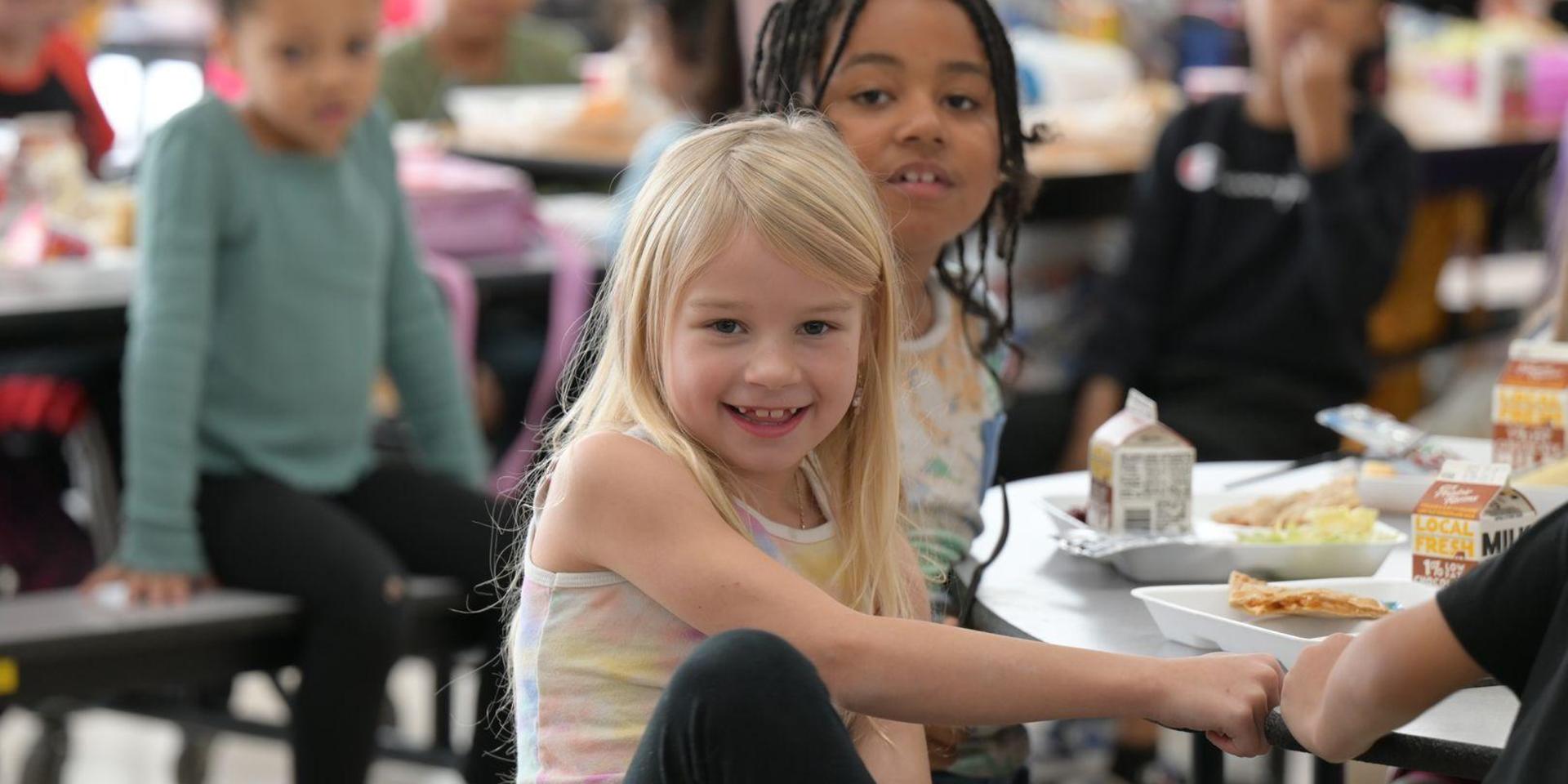 Smiling girl seated at a lunch table with classmates in the background.
