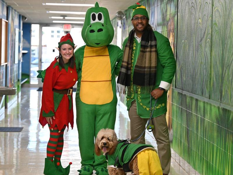 Group of three in costumes, an elf, a dinosaur, and a man with a dog in a school hallway.