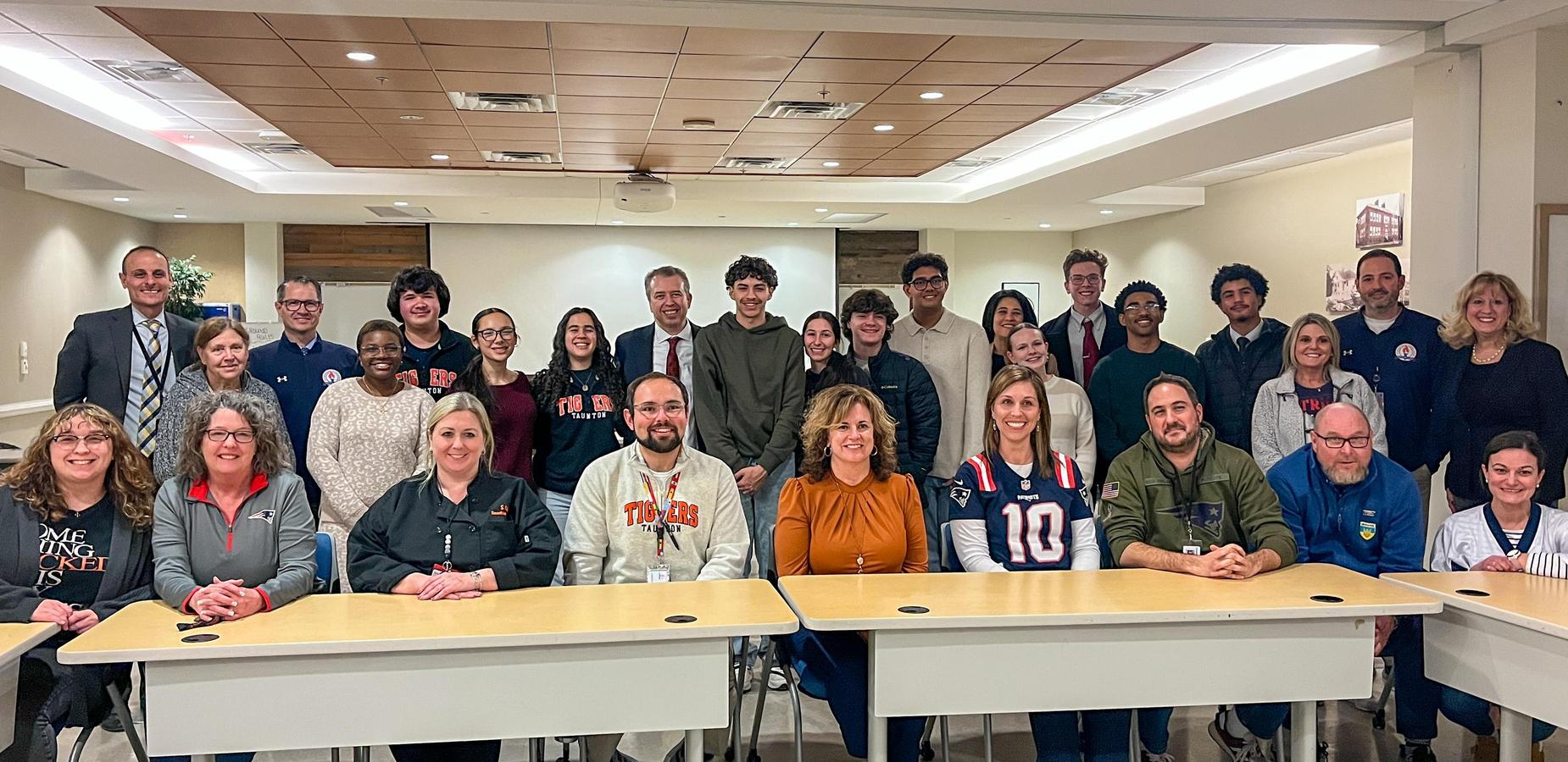 A large group of students and adults sitting and and standing in rows along a long table