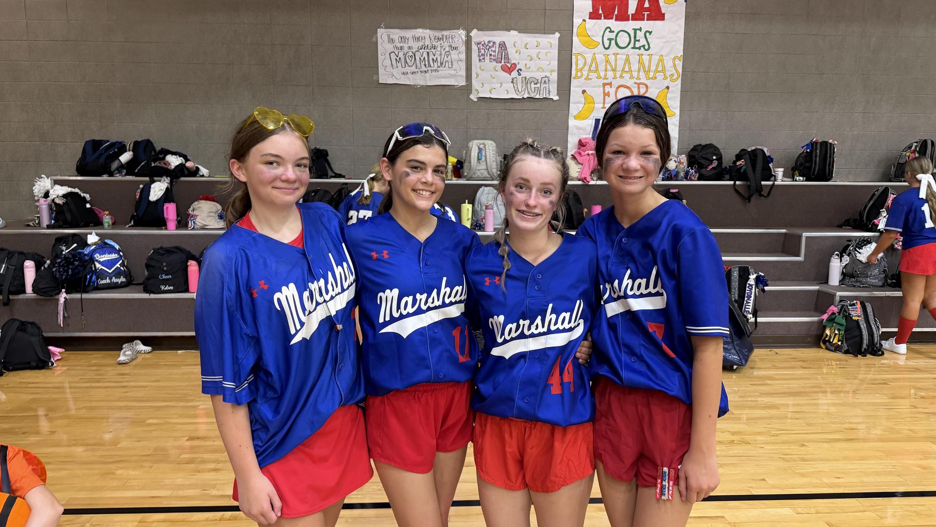 Four girls in sports uniforms smiling together indoors.