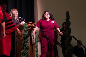 Student in burgundy scrubs walks across the stage to receive her certificate, smiling proudly as a staff member announces her name.