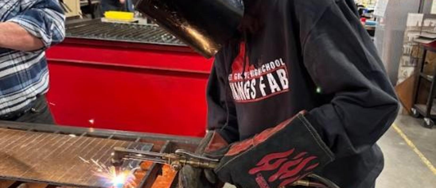 Person wearing welding gloves and a hood, welding a metal piece with sparks flying.
