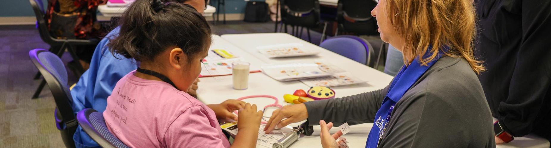 A woman assists a girl with crafts at a table in a classroom setting.