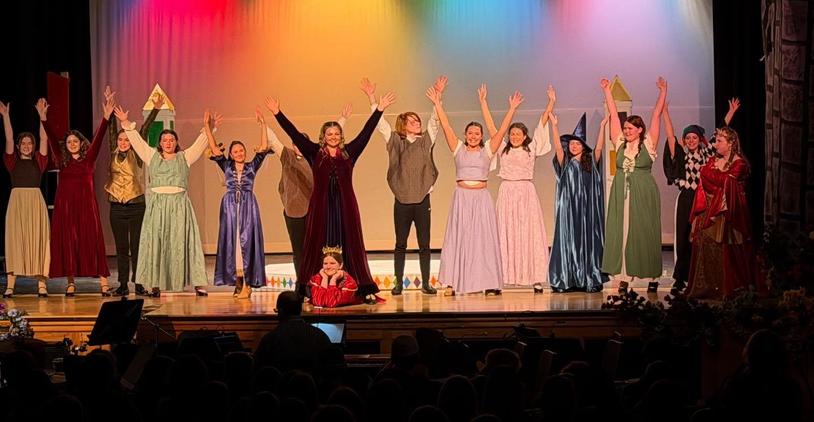 A cast of performers in colorful costumes raising their arms in celebration on stage.