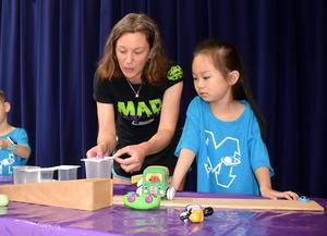 A woman demonstrates an activity to a young girl at a table with colorful materials.